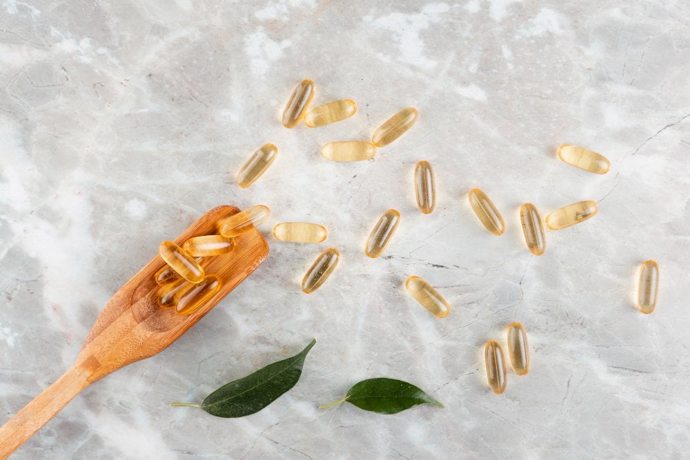 Kapsle omega-3 na světlém mramoru, část na dřevěné lžíci, vedle dva zelené listy