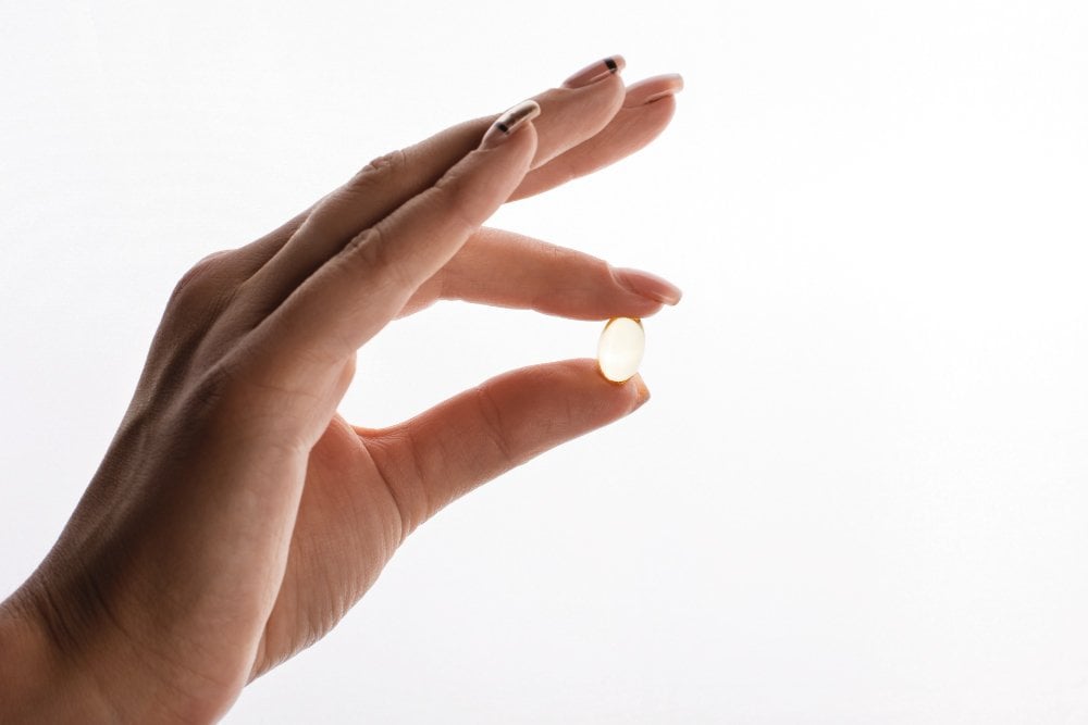 Dlaň drží kapsli omega-3 mezi palcem a ukazováčkem na světlém pozadí